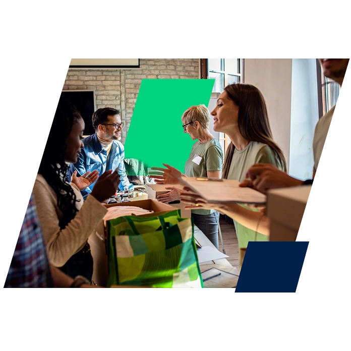 A diverse group of people smiling and collaborating at a community volunteering event. They are standing around tables indoors, organizing items into cardboard boxes and green tote bags, with one person reviewing a clipboard.