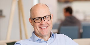 A bald man with glasses and a friendly smile, wearing a light blue button-down shirt. He is posed in a modern, open-plan office with blurred seating and a coworker in the background.