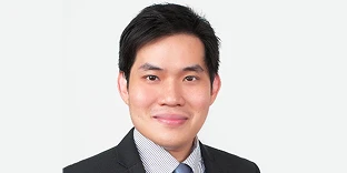 A man with short black hair and a pleasant smile, wearing a black suit jacket over a checkered shirt and a blue tie. He is posed against a solid white background.