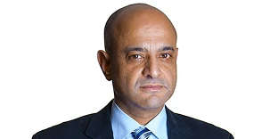 Portrait of a bald man with a serious, professional expression, wearing a dark suit jacket, a light blue shirt, and a striped tie. The background is a solid, neutral white.