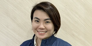 A woman with short dark hair and a warm smile, wearing a navy blue zip-up jacket over a white shirt. She is posed against a background of light-colored, diagonal wooden grain panels.