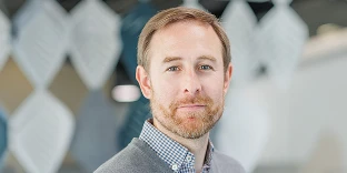 A man with short brown hair and a light beard, wearing a grey sweater over a blue and white checkered shirt. He is looking at the camera with a slight smile in a modern office featuring leaf-shaped wall panels.