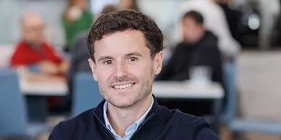 A man with short, curly brown hair and a friendly smile, wearing a navy blue quarter-zip sweater over a light blue collared shirt. He is posed in a blurred, busy office cafeteria or breakroom with colleagues sitting at tables in the background.