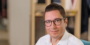 A man with short dark hair and dark-rimmed glasses, wearing a white button-down shirt. He has a slight smile and is posed in a blurred office setting with warm lighting.