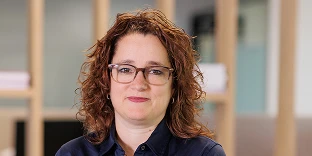 A woman with shoulder-length curly reddish-brown hair and glasses, wearing a dark navy button-down shirt. She has a calm, professional expression and is posed in a blurred office setting with wooden shelving in the background.
