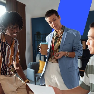 A group of three creative professionals collaborating in a casual office space. A person in a blue blazer holds a coffee cup while discussing a document with a seated colleague, as another coworker listens in.